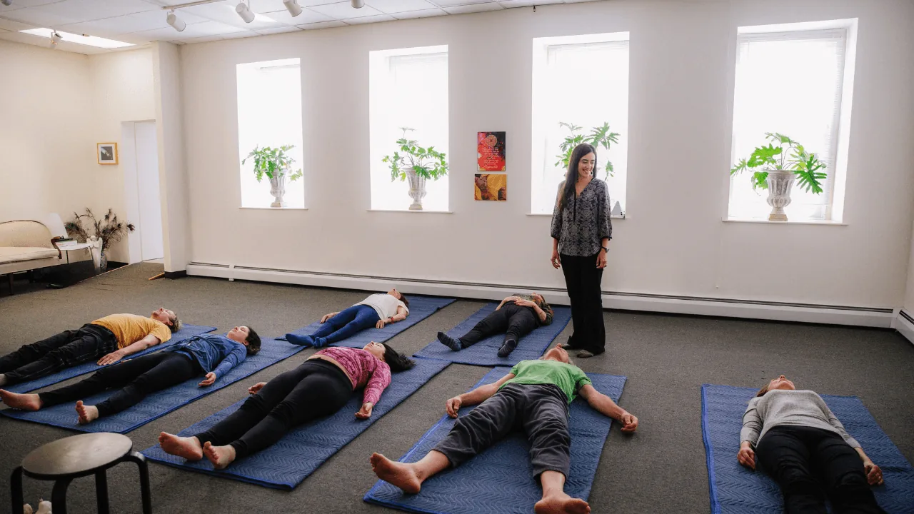 Dayana standing beside participants lying on mats during a Feldenkrais class.